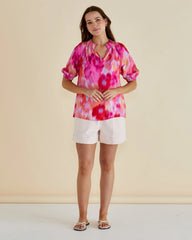 Woman wearing a colorful tie-dye blouse and white shorts against a plain background