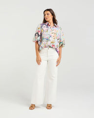 Woman wearing a colorful blouse with a face design and white pants on a white background