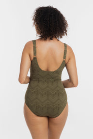 A woman viewed from the back wearing a one-piece swimsuit with a textured geometric pattern in olive green. The swimsuit features thin shoulder straps and a low open back, set against a plain light gray studio background.