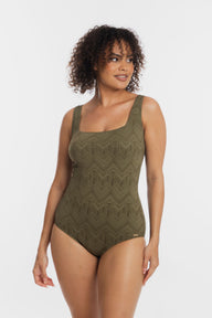 A woman modeling a sleeveless one-piece swimsuit with a textured, geometric chevron-like knit pattern in olive green, set against a plain light gray studio background.