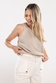 Woman wearing a beige sleeveless knitted top and white pants on a white background