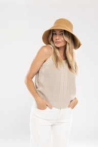Woman wearing a beige sleeveless top and white pants with a tan bucket hat on a white background
