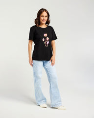 A woman modeling a black short-sleeve t-shirt with a pink and yellow floral graphic on the front, paired with light-wash wide-leg jeans and white sneakers, against a light gray studio background.