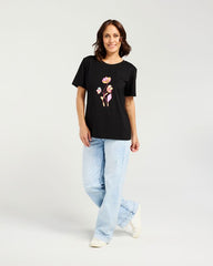 A woman modeling a black short-sleeve t-shirt with a colorful graphic of three cartoon flowers on the front, paired with light-wash jeans and white sneakers, against a plain light gray studio background.