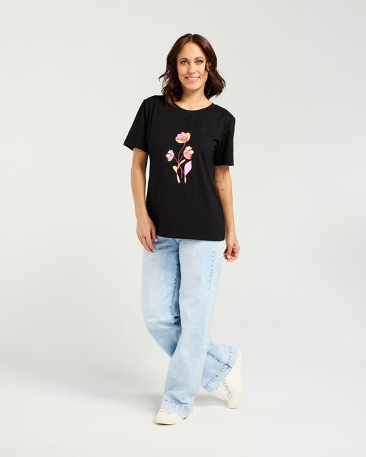 A woman modeling a black short-sleeve t-shirt with a colorful graphic of three cartoon flowers on the front, paired with light-wash jeans and white sneakers, against a plain light gray studio background.