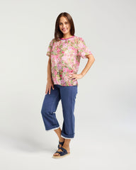 A woman modeling a short-sleeve floral print top in pink, green, and white, paired with blue cropped jeans and navy sandals, against a light gray studio background.