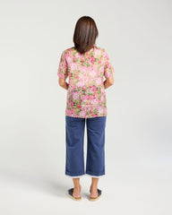 A woman viewed from behind wearing a short-sleeve pink and green floral top with blue cropped pants, standing against a light gray studio background.