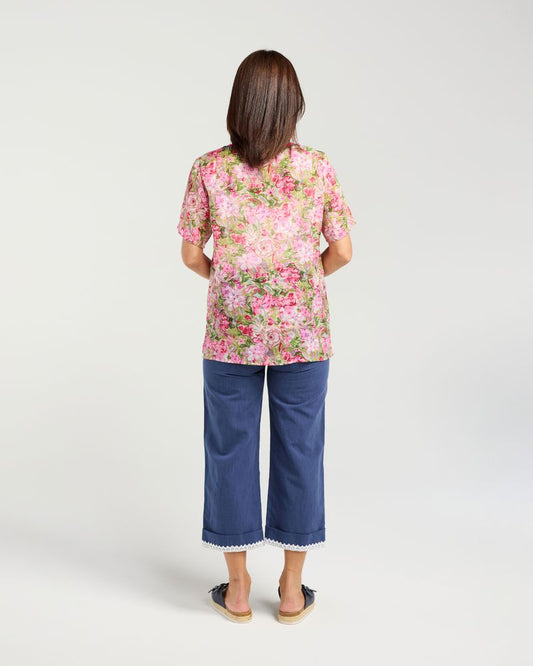 A woman viewed from behind wearing a short-sleeve pink and green floral top with blue cropped pants, standing against a light gray studio background.