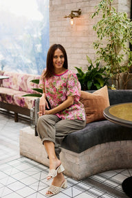 A woman sits on a modern curved sofa in a stylish indoor setting, wearing a pink and green floral short-sleeve top, olive green cropped pants, and white heeled sandals.