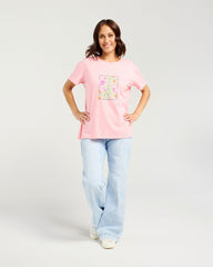 A woman modeling a light pink short-sleeve t-shirt with a colorful floral graphic on the front, paired with light blue jeans and white sneakers, against a light gray studio background.