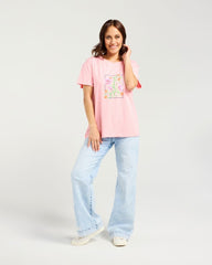A woman modeling a light pink short-sleeve t-shirt with a colorful floral graphic on the front, paired with light-wash wide-leg jeans and white sneakers, against a plain light gray studio background.