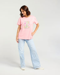 A woman modeling a light pink short-sleeve t-shirt with a colorful floral graphic on the front, paired with light-wash wide-leg jeans and white sneakers, against a light gray studio background.