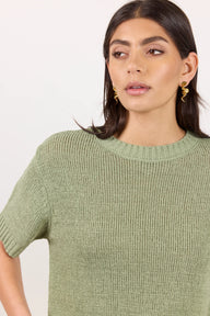 A woman wearing a short-sleeve ribbed knit sweater in sage green, shown from the chest up against a plain light background.