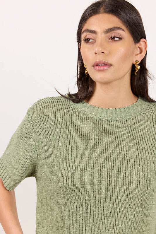 A woman wearing a short-sleeve ribbed knit sweater in sage green, shown from the chest up against a plain light background.