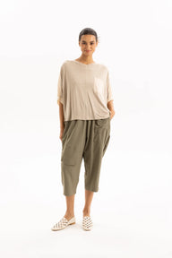 A woman modeling a beige short-sleeve top and olive green cropped pants with white woven shoes, standing against a plain white studio background.