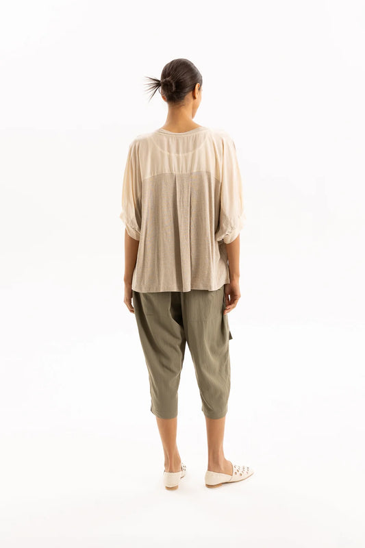 A woman viewed from behind wearing a beige short-sleeve top with a sheer yoke and olive green cropped pants, standing against a plain white studio background.