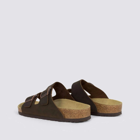 A pair of dark brown double-strap slide sandals with cork-look midsoles, shown from a side angle on a light gray background.