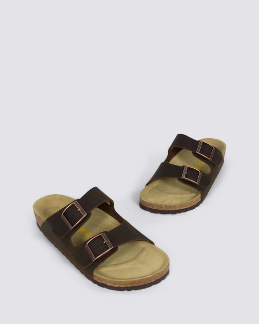 A pair of dark brown double-strap slide sandals with adjustable metal buckles and cork-look midsoles, displayed on a light gray background.
