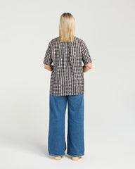 A woman viewed from behind wearing a black and white gingham short-sleeve top and wide-leg blue jeans against a light gray studio background.
