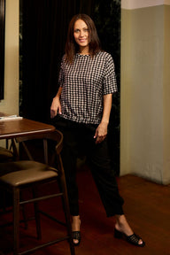 A woman stands indoors wearing a black-and-white gingham short-sleeve top, black cropped pants, and black heeled mule sandals. She is posed beside a wooden table and chair in a dimly lit interior with dark curtains and beige walls.