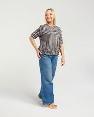 A woman modeling a black and white checkered short-sleeve top with blue flare jeans and beige sandals, standing against a light gray studio background.