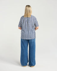 A woman viewed from behind wearing a short-sleeve blue and white gingham check top and wide-leg blue jeans, standing against a light gray studio background.