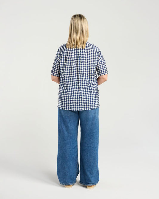 A woman viewed from behind wearing a short-sleeve blue and white gingham check top and wide-leg blue jeans, standing against a light gray studio background.