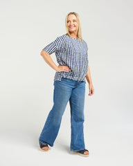 A woman modeling a short-sleeve blue and white gingham check top with blue flare jeans and flat sandals, standing against a light gray studio background.