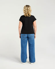 A woman viewed from behind wearing a black short-sleeve t-shirt and wide-leg blue jeans, standing against a light gray studio background.