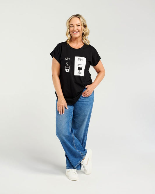A woman modeling a black short-sleeve graphic t-shirt with AM and PM time icons, paired with wide-leg blue jeans and white sneakers, against a light gray studio background.