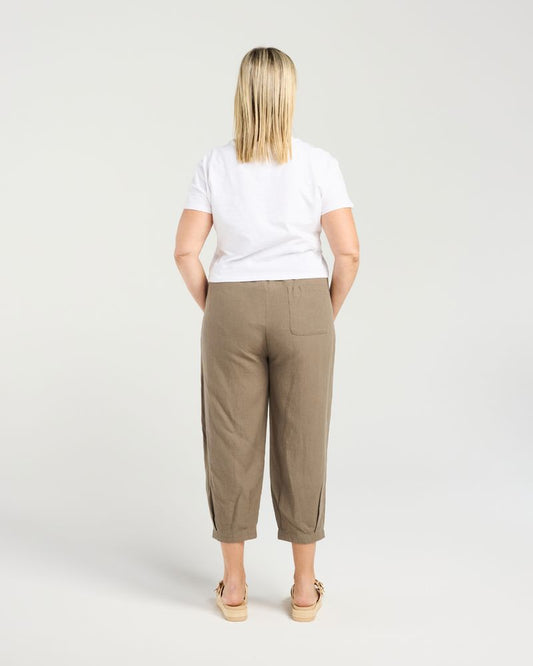 A woman viewed from behind wearing a white t-shirt and cropped khaki trousers, standing against a light gray studio background.