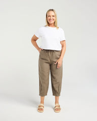A woman modeling a pair of relaxed-fit, cropped ankle-length trousers in a warm taupe-brown color, paired with a white t-shirt and beige sandals, against a light gray studio background.