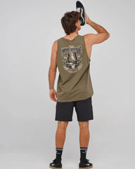 A man shown from the back wearing an olive green sleeveless tank top with a large graphic print on the back, black athletic shorts, and black sneakers, holding a black and white shoe against his head. The background is a plain light gray studio backdrop.