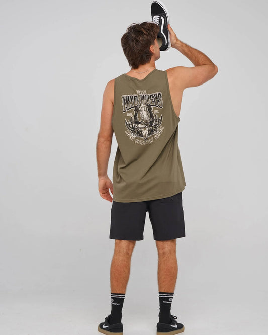 A man shown from the back wearing an olive green sleeveless tank top with a large graphic print on the back, black athletic shorts, and black sneakers, holding a black and white shoe against his head. The background is a plain light gray studio backdrop.
