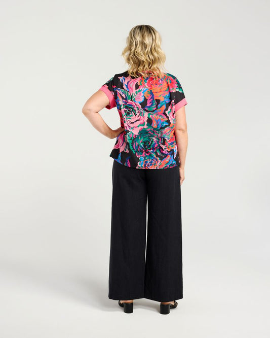 A woman shown from the back wearing a short-sleeve top with a vibrant multicolor floral print and black wide-leg pants, photographed against a light gray studio background.
