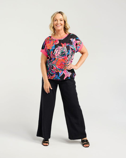 A woman modeling a short-sleeve top with a vibrant multicolor floral print on a black background, paired with black wide-leg pants and black sandals, against a light gray studio background.