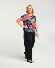 A woman modeling a short-sleeve V-neck top with a vibrant multicolor floral print on a black background, paired with black wide-leg pants and black sandals, against a light gray studio background.