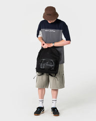 Person wearing a gray and navy shirt with a brown bucket hat, holding a black backpack on a white background.