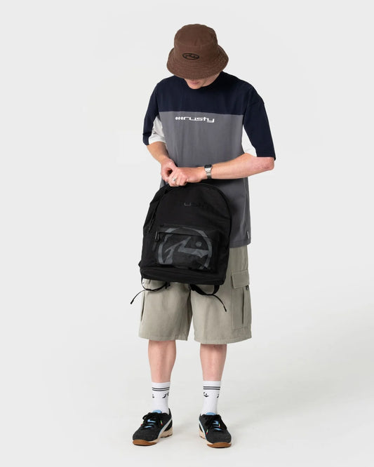 Person wearing a gray and navy shirt with a brown bucket hat, holding a black backpack on a white background.