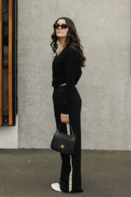 Woman in a black outfit holding a black handbag against a gray wall.