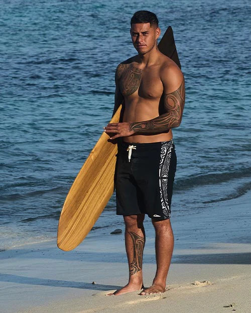 Man with tattoos holding a wooden surfboard on a beach
