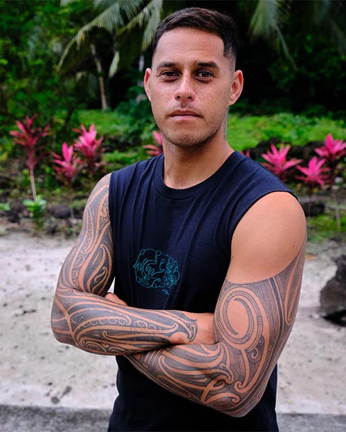 Man with tattoos on his arms standing outdoors with greenery in the background