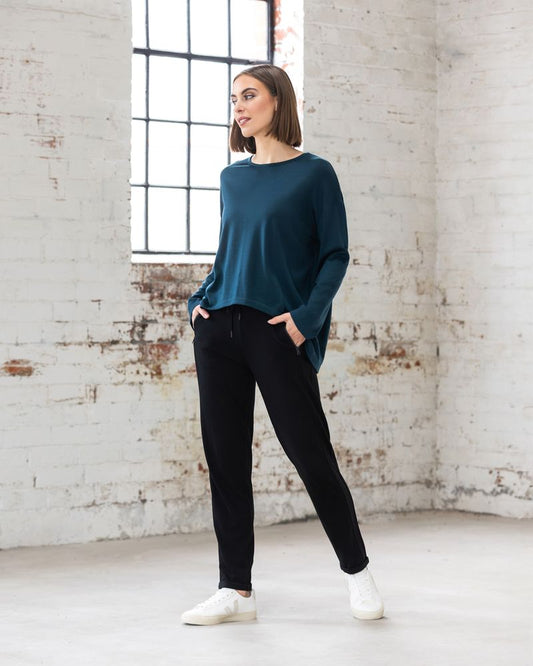 Woman wearing a teal sweater and black pants standing in a room with white brick walls.