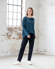 Woman wearing a teal sweater and black pants standing in a room with white brick walls.