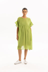 A woman modeling a loose, short-sleeved midi dress in a light olive green color, paired with white sneakers, against a plain white studio background.