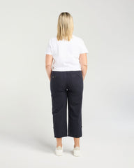 A woman viewed from behind wearing dark navy cropped pants, a white t-shirt, and white sneakers against a light gray studio background.