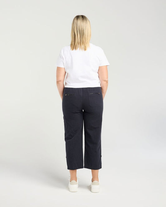 A woman viewed from behind wearing dark navy cropped pants, a white t-shirt, and white sneakers against a light gray studio background.