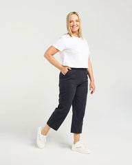 A woman modeling a white short-sleeve crewneck t-shirt and dark navy cropped trousers, paired with white sneakers, against a light gray studio background.
