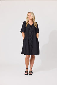 A woman modeling a black short-sleeve button-front dress with side pockets, standing against a plain light gray studio background.