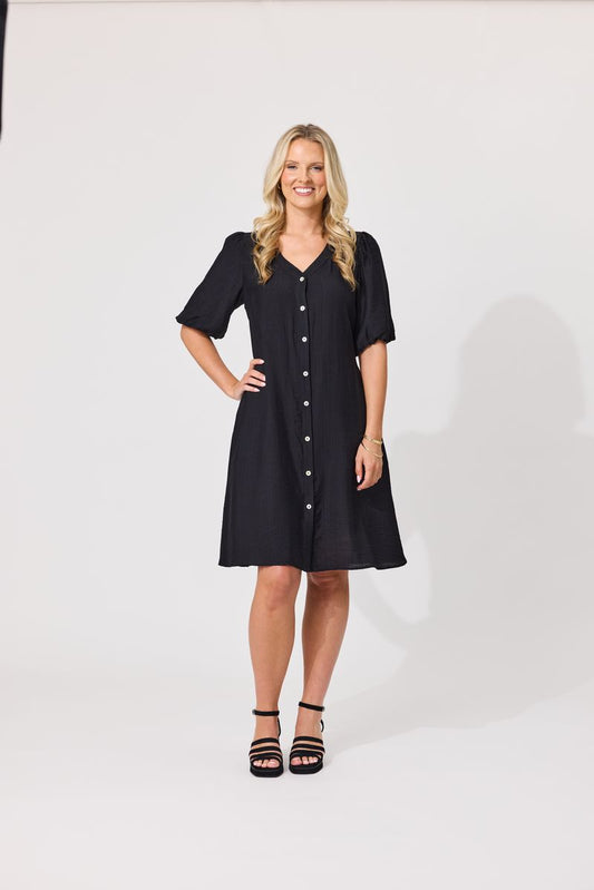 A woman modeling a black short-sleeve button-front dress with a V-neck and flared skirt, standing against a plain light gray studio background.